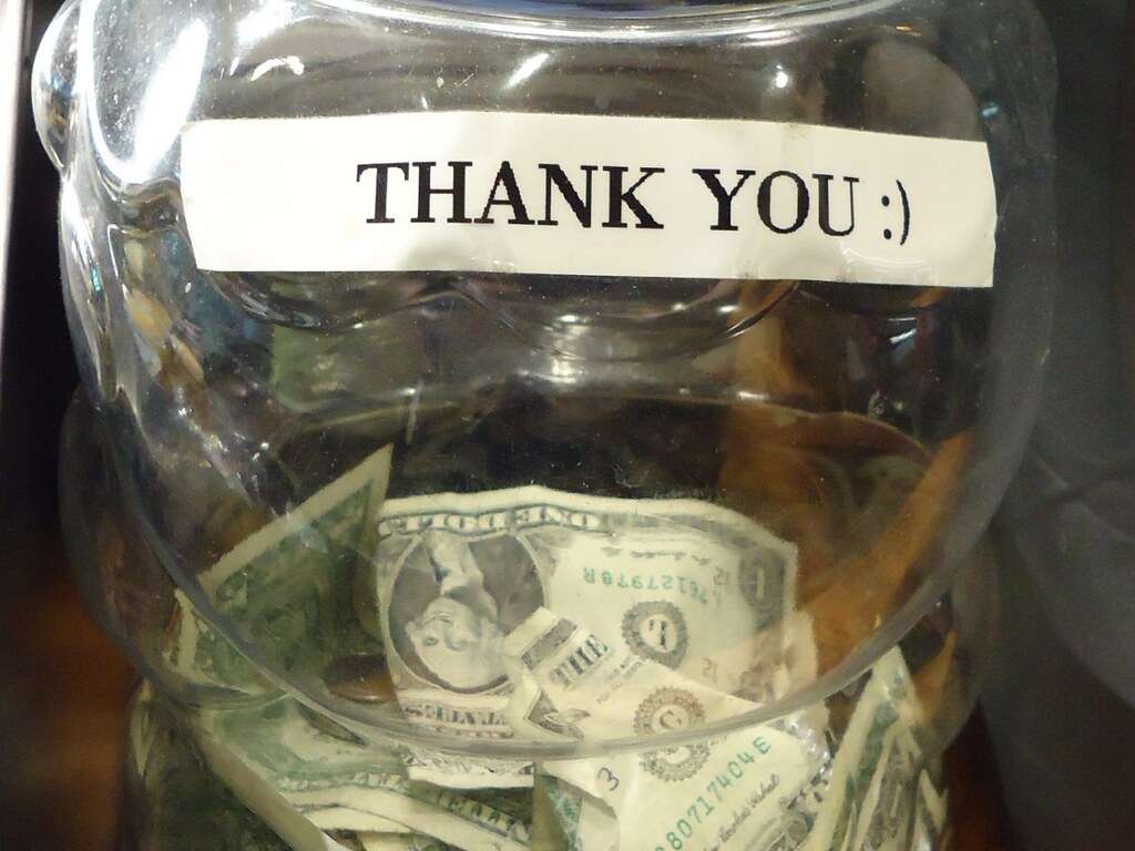 A clear plastic tip jar with a white label that reads "THANK YOU :)" and a one-dollar bill behind the label, standing on a wooden counter in a restaurant in New Jersey